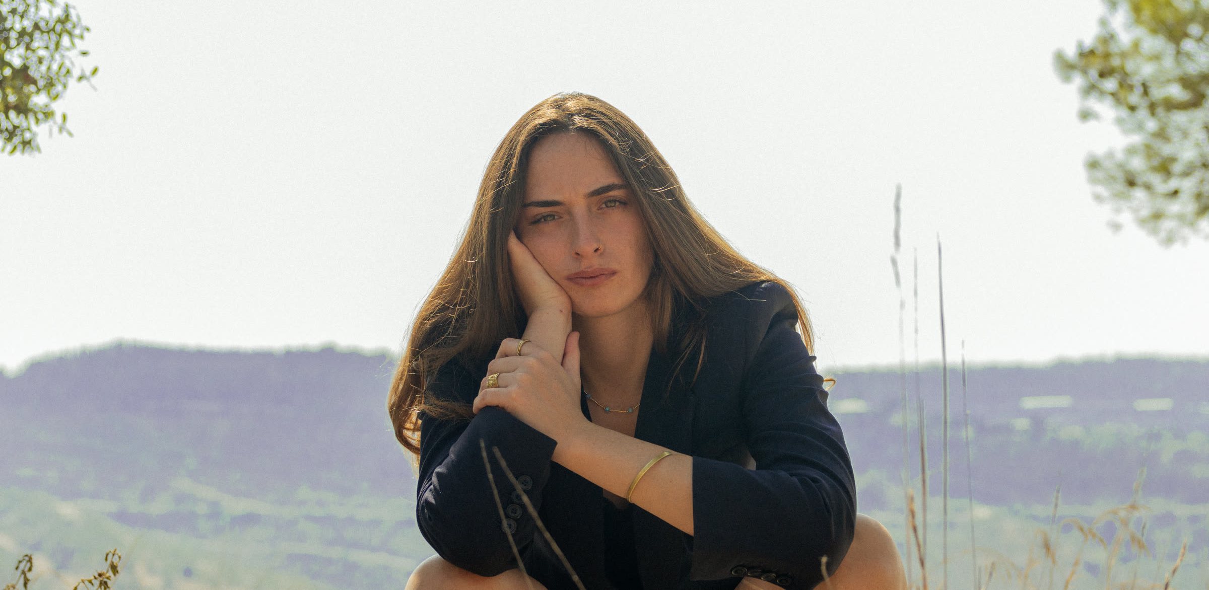 Rivka Braverman seated on a hillside, looking toward the camera.